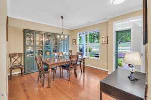 A dining room with a wooden table and six chairs, a green cabinet displaying dishes, and a candle centerpiece. Large window with a view of a fenced yard and trees. Hardwood floor, beige walls, white trim, and a lit chandelier.