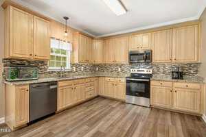 A modern kitchen features wooden cabinets, a stainless steel stove, dishwasher, and a coffee maker. The countertop is granite, and a fish tank is next to the sink under a window. The backsplash has a mosaic tile design, and the floor is wood.