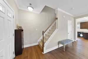 A hallway with a wooden staircase, beige walls, and a small decorative table. A closed door is on the left, leading to an open kitchen area. A framed picture rests on a dark cabinet beneath a ceiling light.