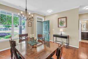 Dining room with wooden table and six chairs. A chandelier hangs overhead, and a table centerpiece with candles is present. The room has large windows, an open door, artwork on beige walls, and a side table with a lamp.
