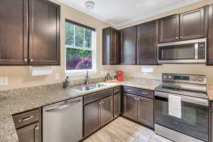 Modern kitchen with dark wooden cabinets, granite countertops, stainless steel appliances including a stove and dishwasher, a microwave, and a window overlooking greenery. Light-colored tile flooring and a simple, clean design.