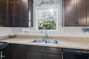A kitchen sink with a double basin is centered under a window with sunlight streaming in. The countertop is beige with a speckled pattern, and dark wooden cabinets surround it. A paper towel holder and a small jar are positioned nearby.