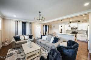 A contemporary living room features a gray sofa and two navy chairs around a light wood coffee table on a patterned rug. The open kitchen has white cabinets, stainless steel appliances, and two pendant lights over a breakfast bar.