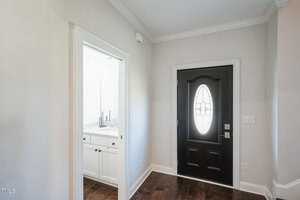 A hallway with a dark front door featuring an oval glass window. To the left is an open doorway leading to a bathroom, visible with a sink and faucets. The flooring is dark wood, and the walls are painted light gray.