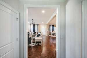 View from a hallway into a spacious living room with wooden floors. The room features a seating area with sofas, a chandelier, and large windows with curtains. Adjacent is a dining area with a table and chairs. Walls are painted in neutral tones.