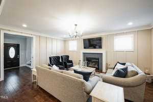 A cozy living room with a modern chandelier, two comfortable gray sofas, black armchairs, and a fireplace. A flat-screen TV is mounted above the fireplace. The room features wooden flooring and a circular coffee table at the center.