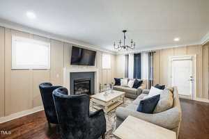 A cozy living room with beige walls and a wooden floor. It features a fireplace, a wall-mounted TV, blue and gray sofas, a coffee table, and a chandelier. Natural light streams in through windows and a glass door.