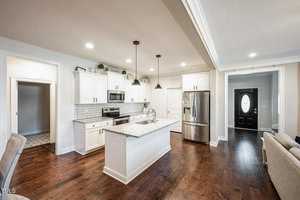 A modern kitchen with white cabinets, stainless steel appliances, and a center island with a granite countertop. Dark hardwood floors complement the space, and pendant lights hang above the island. A black door is visible in the background.