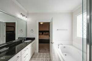 A modern bathroom with a black granite double sink vanity, large mirror, and bright lighting. There's a bathtub with a window above it, and an open door leading to a bedroom with a bed in view. The floor features patterned tiles.