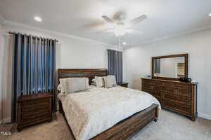 A cozy bedroom with a large wooden bed, ornate headboard, and light-colored bedding. Flanked by two wooden nightstands, it features a dresser with a mirror. Gray curtains cover the windows, and a ceiling fan is above. The carpet is beige.