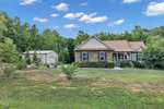 A small house at 3723 Summer Springs Drive in Franklinton features a stone and siding exterior, nestled in a grassy yard surrounded by trees. A detached garage stands to the left. The sky is blue with clouds, and a United States flag flutters proudly by the entrance.