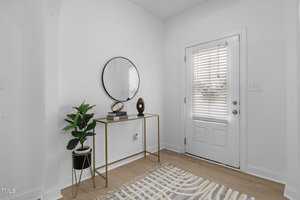 A minimalist entryway with a white door and blinds. A small table with a gold frame holds a potted plant, a round mirror, a decorative sculpture, and a book. A patterned rug covers the light wood floor.