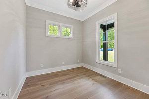 An empty room with light gray walls, white trim, and a wooden floor. It features two windows on adjacent walls, allowing natural light. A modern ceiling fan is centered above. A partial view of a blue car and green grass is visible outside.