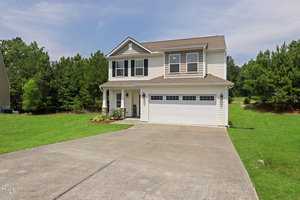 A two-story house with beige siding and dark shutters, featuring a double garage and a neatly maintained lawn. Surrounded by trees, the home sits under a blue sky with a concrete driveway leading up to it.