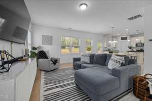 A modern living room with a gray sectional sofa, striped rug, and a wall-mounted TV. Large windows let in natural light. A dining area with a table and chairs is in the background, adjacent to a kitchen with white cabinets and stainless steel appliances.