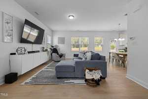 A modern living room with a large wall-mounted TV, white cabinets, and gray accent chairs. A gray sectional sofa is centered on a striped rug. Light pours in from windows, and a view of a dining area is visible in the background.