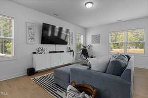 A modern living room with a large wall-mounted TV above a sleek white console. A gray sectional sofa faces the TV, adorned with cushions. Light floods the room through several windows, and various decorative items are neatly arranged.