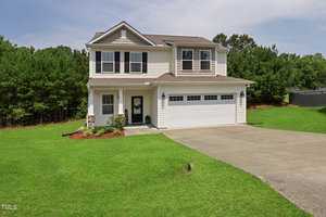A two-story suburban house with beige siding and white trim, featuring a two-car garage and a well-maintained lawn. The house is bordered by a lush green hedge of trees, and the driveway curves gently to the right.