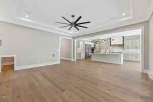 Spacious modern room with light wood flooring. Features an open-concept kitchen with a large island and pendant lights, white cabinetry, and stainless steel appliances. A ceiling fan hangs in the center of the living area. Walls are painted light gray.