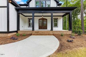 A modern two-story house with a large wooden front door, black-trimmed windows, and a spacious porch. The exterior features white siding and brick accents. There is a curved concrete pathway leading to the entrance, surrounded by minimal landscaping.