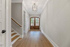 A bright hallway with light wood flooring and neutral-colored walls featuring a double wooden front door with arched windows. A modern light fixture hangs from the ceiling. A staircase with a wooden handrail is visible on the left.