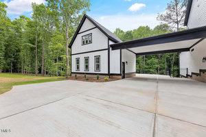 A modern house with white siding and black trim, featuring tall windows and a covered driveway. It is surrounded by lush green trees and has a spacious concrete driveway under a clear blue sky.