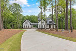 A modern two-story white house with black trim, located at 984 Browning Place in Youngsville, is surrounded by tall trees. A long concrete driveway leads to the front, and the neatly manicured lawn complements the lush green forest under a partly cloudy sky.