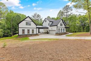 A modern farmhouse with a white exterior, black trim, and a dark roof is surrounded by trees and greenery. The house features large windows, a spacious driveway, and an adjoining garage under a clear blue sky.