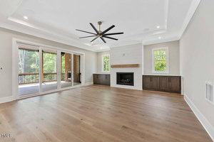 Spacious living room with light wooden floors, white walls, and a ceiling fan. Features a fireplace flanked by small windows and built-in cabinets. Large sliding glass doors open to a covered deck with a view of trees.