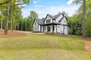 A large, modern white house with black trim sits in a lush, wooded area. The house features multiple gables and large windows. A manicured lawn extends across the foreground, surrounded by tall trees under a clear blue sky.