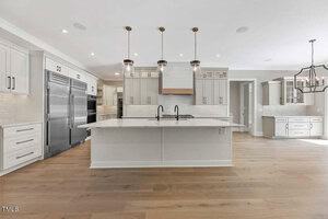 A spacious modern kitchen with white cabinetry, a large island with a sink, and three pendant lights above. Stainless steel appliances are on the left, hardwood floors throughout, and a dining area visible in the background.