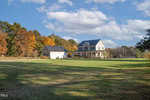 A charming two-story farmhouse located at 3600 Trappers Run in Wake Forest features a large porch that beckons you to relax and enjoy the view. Set in a grassy field, the property includes a detached garage and is surrounded by trees displaying vibrant autumn colors under a sky with scattered clouds.
