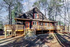 A rustic, dark wooden house with red window shutters, surrounded by tall trees. The front features a wide wooden porch with railings and a central staircase leading to the entrance. The setting is serene and wooded, under a clear blue sky.
