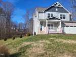 A two-story gray house with white trim at 3447 Lilac Lane features a covered porch and a garage, nestled on a grassy slope in Wake Forest. Small shrubs and trees surround the home, while the clear blue sky indicates a sunny day.