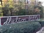 A large wooden sign with the words "Shelton Hills" written in cursive is surrounded by greenery and bushes at 1556 Dogwood Drive. Trees with autumn leaves create an enchanting backdrop in Oxford.