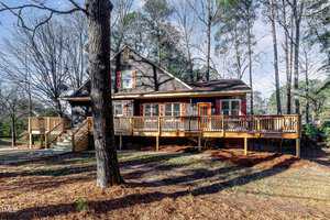 A rustic wooden house surrounded by tall trees and a spacious deck. The exterior features brown siding with red shutters. The yard is covered with pine needles and bathed in sunlight on a clear day.
