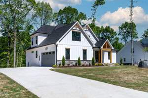 A modern, two-story white house with a dark roof and wooden accents. It features large windows, a stone base, and a spacious driveway leading to a two-car garage. The surrounding area is landscaped with trees and a green lawn.
