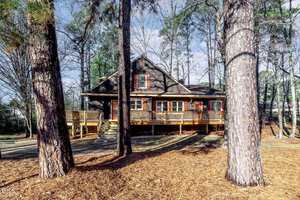 A rustic, dark brown, two-story wooden cabin with a large front porch is surrounded by tall pine trees and a forest floor covered in pine needles. The sky is partly cloudy, casting soft light on the scene.