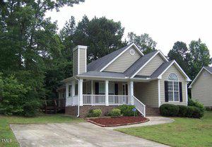 A charming single-story house with light brown siding and white trim, featuring a front porch with columns. A neatly landscaped front yard and a concrete driveway lead up to the home. Lush trees surround the property, adding a sense of seclusion.