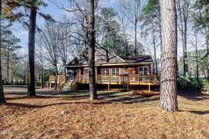 A rustic wooden house with a wraparound porch surrounded by tall pine trees. The yard is covered in brown pine needles and the sky is partly cloudy. The setting conveys a serene and natural ambiance.