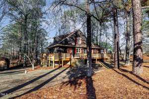 A rustic wooden cabin surrounded by tall pine trees in a forest setting. The cabin features a large wraparound deck and is set on a sloped, pine needle-covered ground. The sky above is clear and blue, suggesting a sunny day.