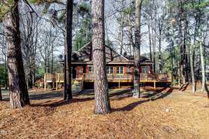A rustic cabin surrounded by tall pine trees with a large wooden deck. The ground is covered in pine needles, and the trees' shadows stretch across the scene, creating a tranquil forest atmosphere.