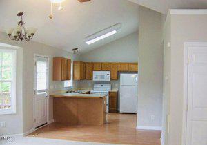 A bright kitchen with light wood cabinets, white appliances, and a vaulted ceiling. Natural light enters through a window and a door with glass panels. A hanging light fixture is visible above the open space. The floor is hardwood.