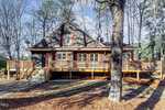 A rustic brown cabin on Creedmoor with a large wooden deck, surrounded by tall trees. The front features red shutters on windows and a wooden staircase leading to the entrance. It's set against a backdrop of bare winter trees and scattered pine needles.