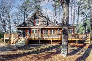 A rustic brown cabin on Creedmoor with a large wooden deck, surrounded by tall trees. The front features red shutters on windows and a wooden staircase leading to the entrance. It's set against a backdrop of bare winter trees and scattered pine needles.