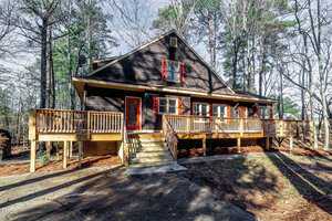 A rustic, two-story cabin with dark wooden siding, red trim, and red shutters is surrounded by tall pine trees. It features a large wooden deck with stairs leading down to a concrete path amidst a wooded area.