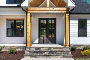 A modern house entrance featuring a wooden porch with a peaked roof, dark double doors, and white siding. The stone steps are bordered by small shrubs, and sunlight creates shadows on the structure.
