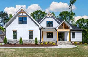 Located at 995 Browning Place in Youngsville, this modern suburban house features a white exterior with stone accents and black-framed windows. It boasts a gabled roof, a covered entryway with wooden beams, and is complemented by a well-maintained front lawn against a backdrop of trees.