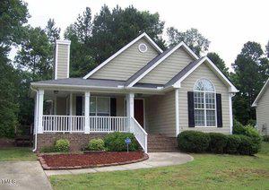 A charming, single-story house on 2777 Clifton Avenue in Creedmoor boasts beige siding, white trim, and a welcoming front porch. It features a gable roof, chimney, and arched window. A short brick walkway leads to the entrance surrounded by lush shrubs and trees.