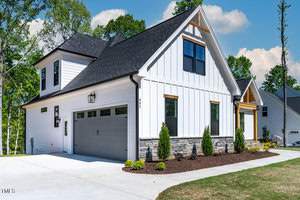 A modern white house with a dark gray roof, featuring a spacious garage, large windows, and a gable roof. The front yard is landscaped with small shrubs and a neat driveway. Trees and a blue sky are visible in the background.